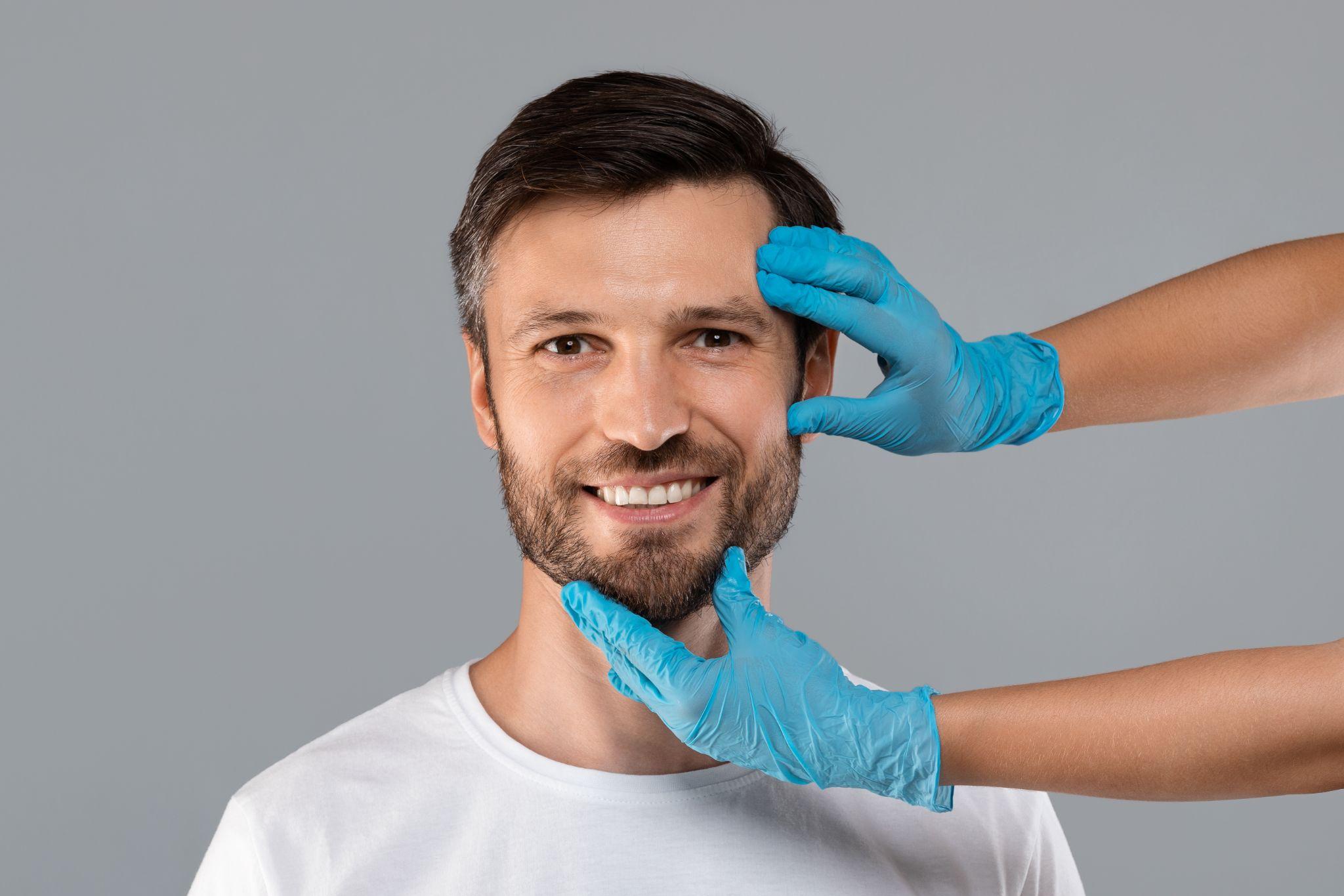 Cosmetician hands in protective medical gloves touching smiling man face over grey studio background. Plastic surgery and aesthetic cosmetology concept.